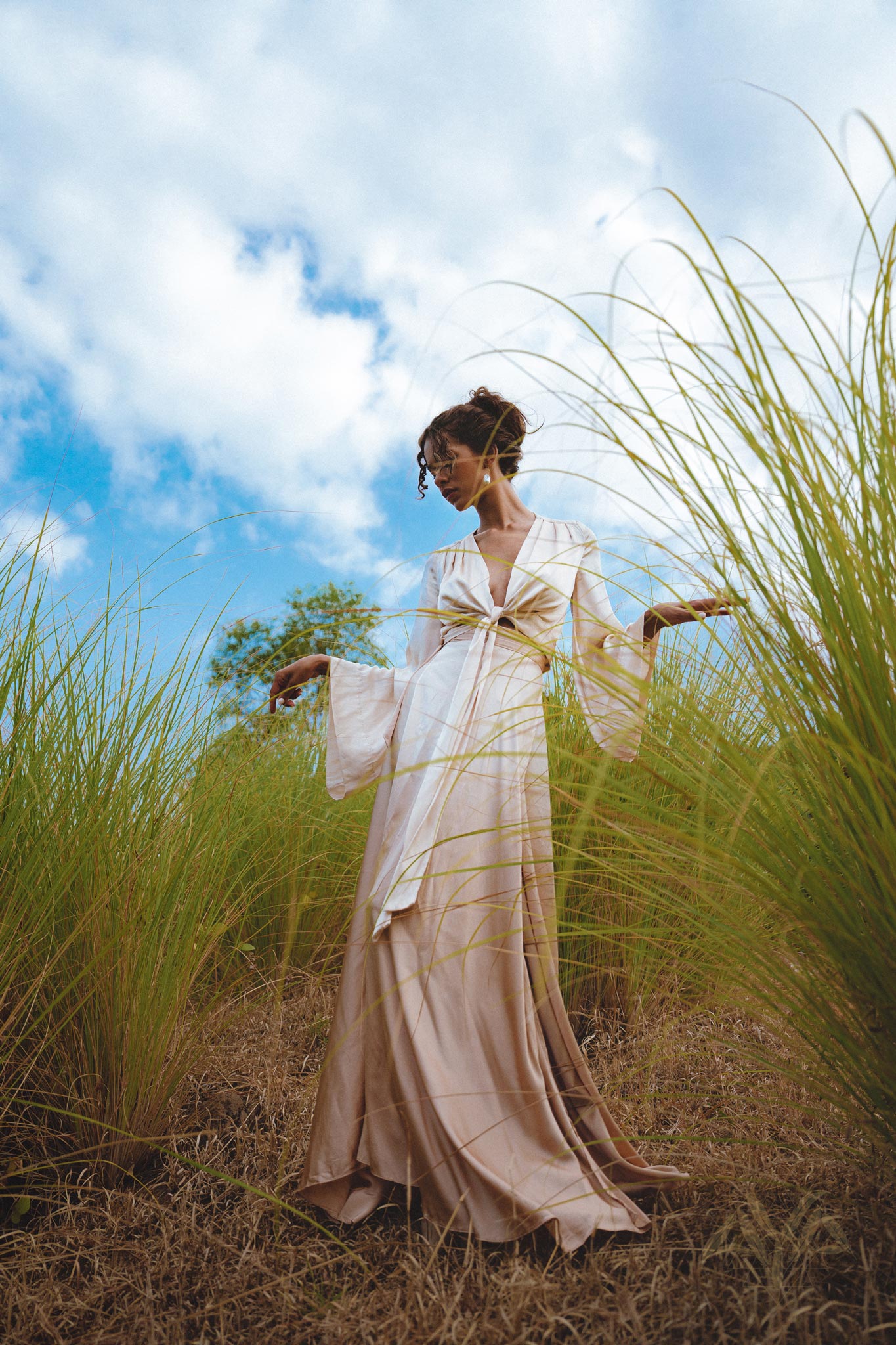 Model wearing a Powder Pink Venus Peace Silk Skirt, a minimalist wrap skirt made from organic materials, standing gracefully in a field of tall grass under a bright blue sky. The skirt flows elegantly as she poses with her arms raised.