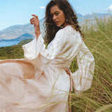 A model poses gracefully in a field, wearing the Powder Pink Venus Peace Silk Skirt, a minimalist wrap skirt that flows elegantly with her movements. The skirt's organic fabric complements the natural landscape, featuring lush grass and a scenic backdrop. The soft pastel hue and stylish design make it a perfect choice for a summer outfit.