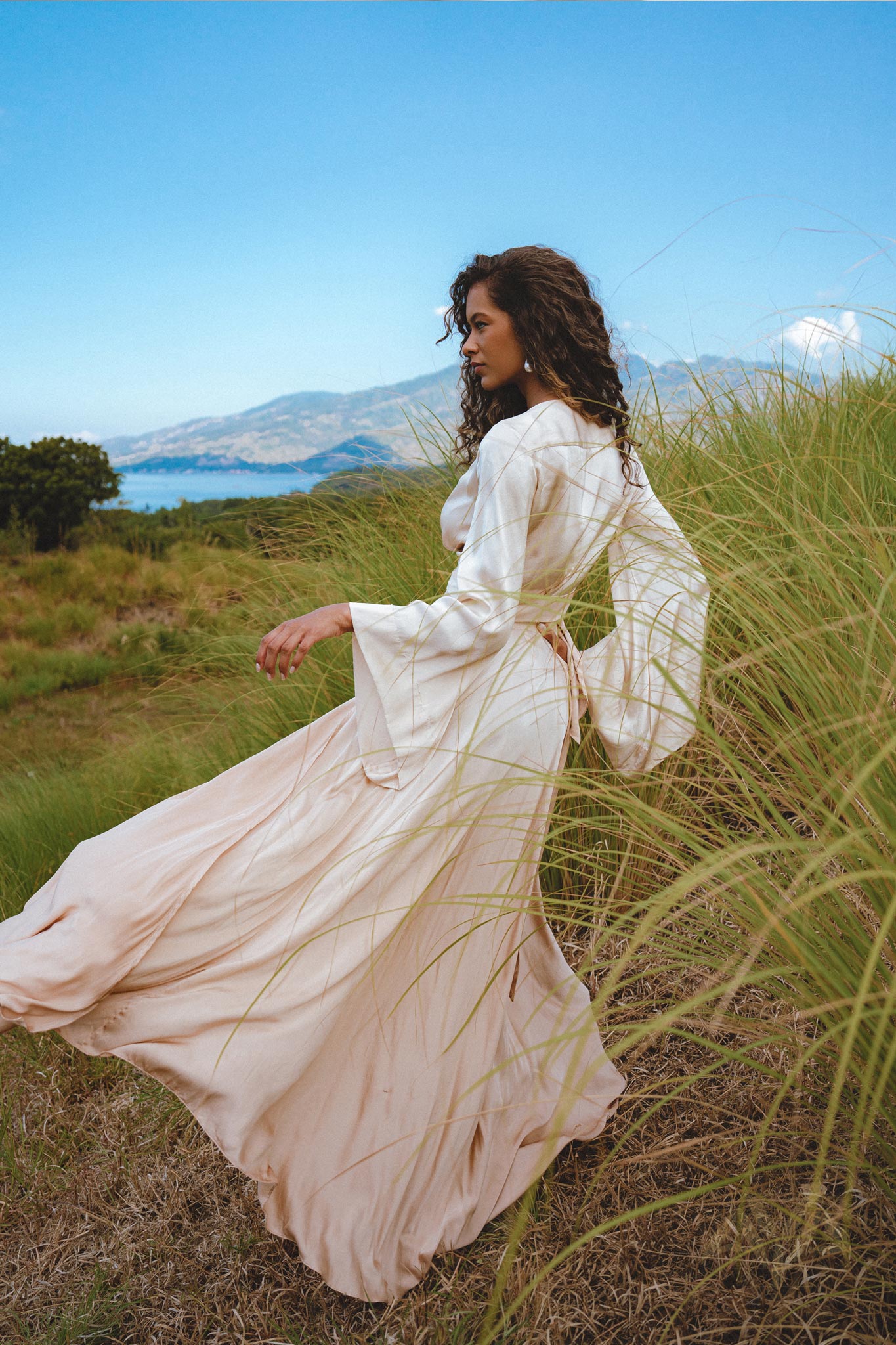 model wearing a Powder Pink Venus Peace Silk Skirt, showcasing a minimalist wrap skirt design, standing in a lush green field with mountains and a body of water in the background. The skirt flows elegantly with a gentle breeze, highlighting its organic summer aesthetic.