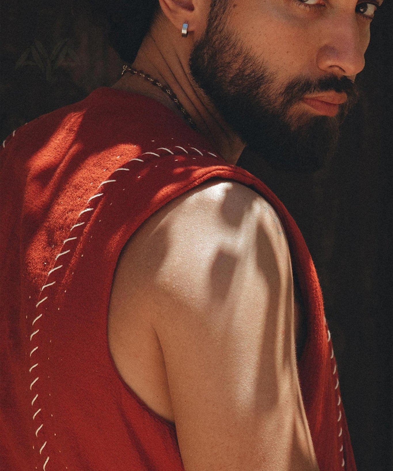 A man with a beard and an earring looks over his shoulder, wearing an AYA Sacred Wear Red Hand Embroidered Vest for Men. The lighting casts shadows on his skin, highlighting his muscular arm and intense gaze.