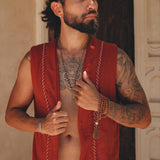 A person with long, dark hair tied back is wearing a Red Hand Embroidered Vest for Men from AYA Sacred Wear and white pants, standing in front of an ornate wall. They are adorned with tattoos on their arms and chest and are holding a beaded bracelet in one hand.