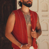 A man with a beard stands confidently in front of a carved wooden door, wearing an AYA Sacred Wear Red Hand Embroidered Vest for Men and white pants, showcasing necklaces and tattoos.