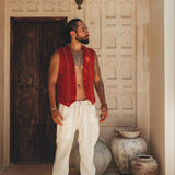 A man stands in front of an ornate wooden door, clad in a vibrant Red Hand Embroidered Vest by AYA Sacred Wear and white pants. He is barefoot and surrounded by earthen pots, as sunlight filters through the leaves, creating intricate patterns on the wall.