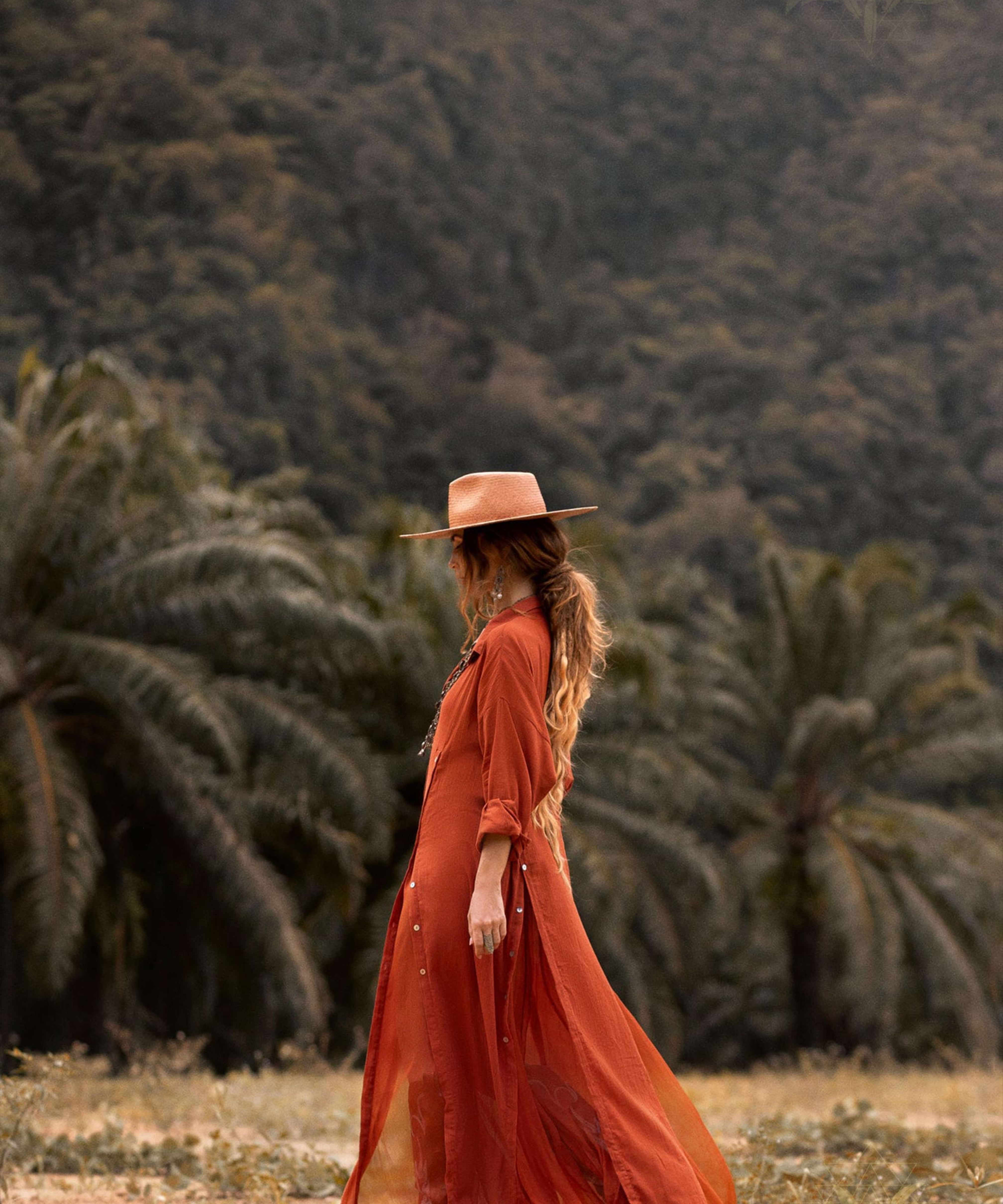 A woman stands contemplatively in front of palm trees and forested hills, wearing a flowing Red Kannika Shirt Dress by AYA Sacred Wear. Her long, colorful hair blends perfectly with the natural backdrop, complementing her wide-brimmed hat.