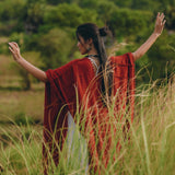 Amidst the lush, green landscape, a person stands with outstretched arms in tall grass. They wear the Red Poncho Robe by AYA Sacred Wear, a flowing bohemian garment that resembles a poncho cape. Their long dark hair is tied up, complementing the essence of traditional crafts that Bali is renowned for.