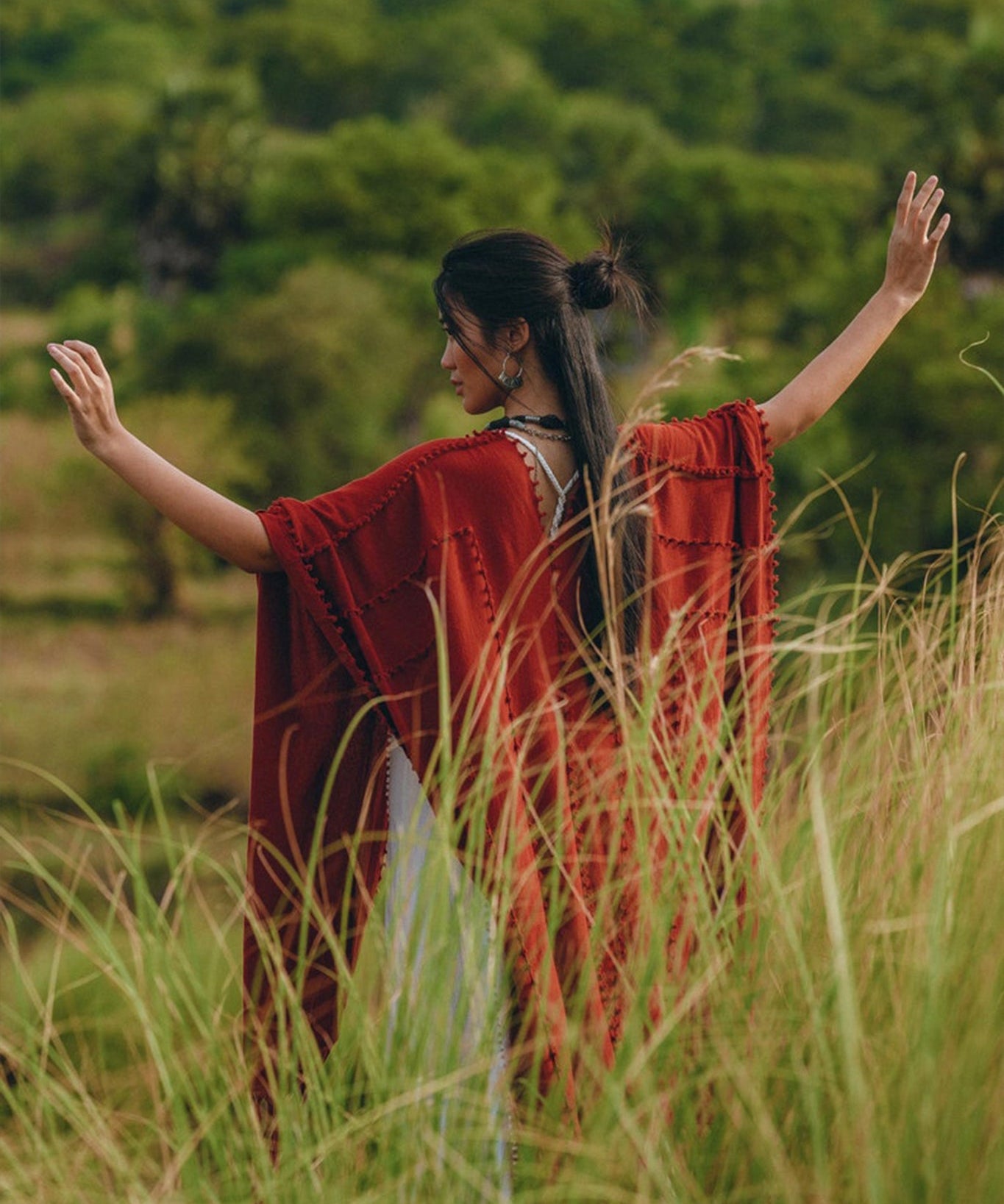 Amidst the lush, green landscape, a person stands with outstretched arms in tall grass. They wear the Red Poncho Robe by AYA Sacred Wear, a flowing bohemian garment that resembles a poncho cape. Their long dark hair is tied up, complementing the essence of traditional crafts that Bali is renowned for.