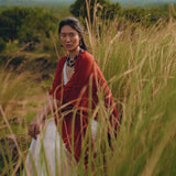 Draped in an AYA Sacred Wear Red Poncho Robe, a woman wearing a Boho Cape and white dress sits amidst tall grass in a serene, lush landscape with hills in the background. The atmosphere is calm and contemplative, evoking a connection to traditional crafts.