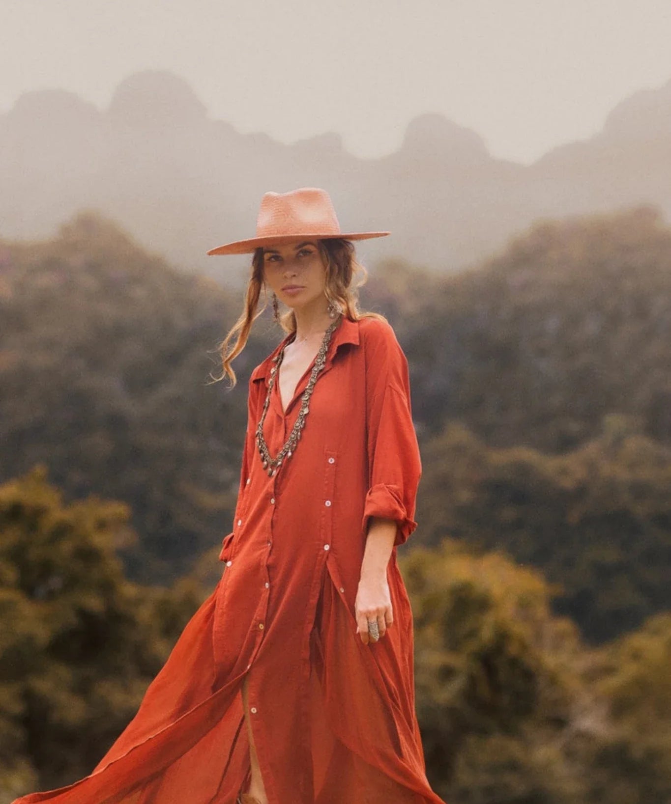 A woman outdoors wears the AYA Sacred Wear Red Kannika Shirt Dress—an adjustable, multiway button-down kaftan—paired with a wide red hat and layered jewelry. Misty trees and mountains blur in the background.