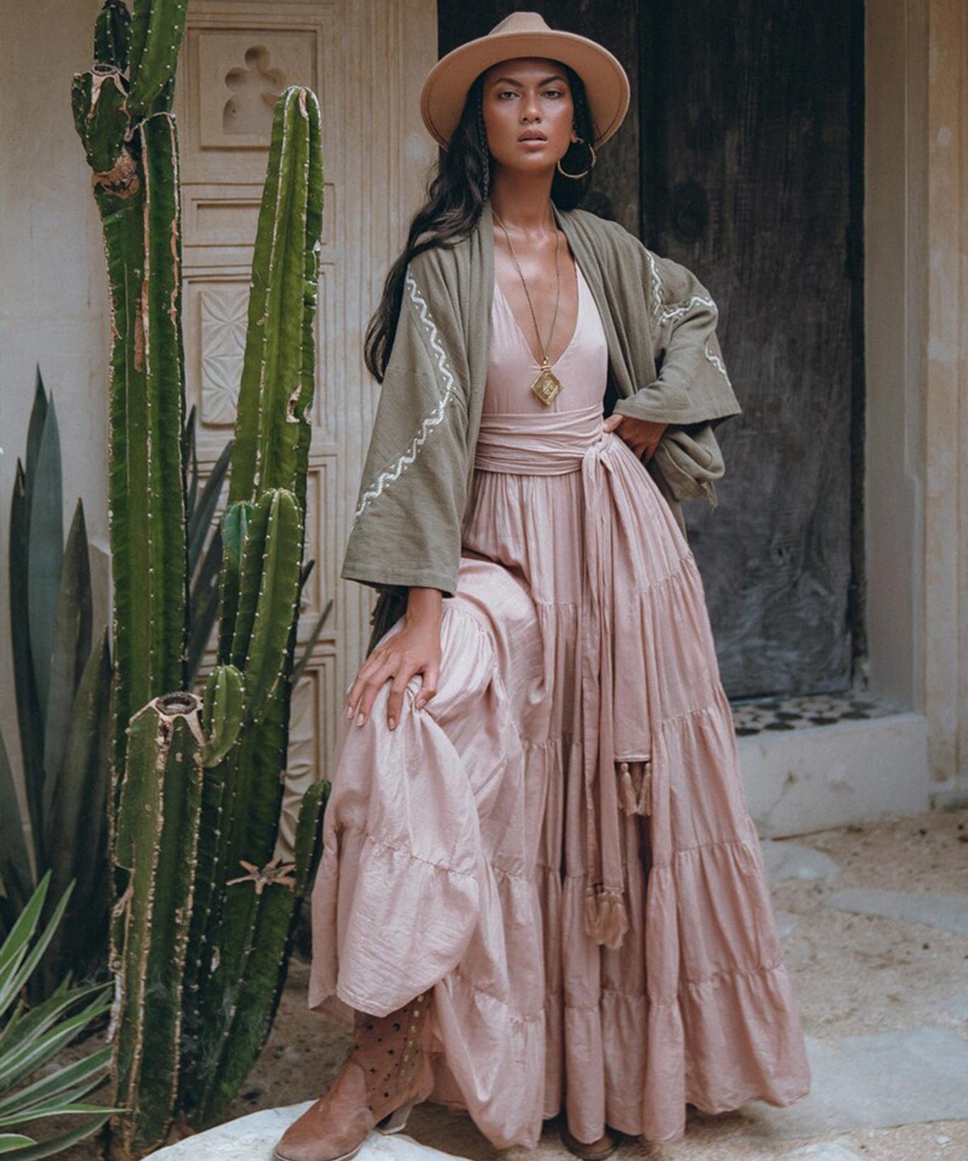 A woman wearing a wide-brimmed hat and pink maxi dress stands beside a towering cactus. She confidently poses in front of a rustic wooden door in the desert, draped in an AYA Sacred Wear Sage Colour Organic Cotton Poncho Cape, paired with brown boots.