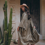 A person wearing the AYA Sacred Wear Sage Colour Organic Cotton Poncho Cape, featuring a bohemian design, stands beside tall cacti in front of a richly carved wooden door. The setting exudes a rustic and bohemian vibe.