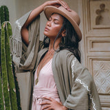 A woman wearing the Sage Colour Organic Cotton Poncho Cape by AYA Sacred Wear stands serenely with her eyes closed. She is dressed in a pink dress adorned with shamanic embroidery, complemented by a tan hat and large hoop earrings. Her green shawl, draped over the bohemian cardigan, adds to her tranquil aura as she leans against a tall cactus in a rustic setting with intricately carved wooden doors, gently placing one hand on her hat.