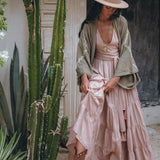A woman stands beside a tall cactus, gazing downward, adorned in an AYA Sacred Wear ensemble: a tan hat and Sage Colour Organic Cotton Poncho Cape with shamanic embroidery. Her flowing pink dress is complemented by a pendant and brown boots, perfectly capturing her bohemian style.