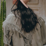 A woman with long dark hair, wearing the AYA Sacred Wear wide-brimmed pink hat and a Sage Colour Organic Cotton Poncho Cape featuring bohemian shamanic embroidery, stands with her back to the camera. A tall cactus is beside her, and a decorative wall serves as the backdrop.