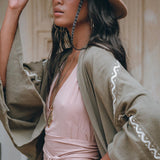 A person wearing a beige hat and an AYA Sacred Wear Sage Colour Organic Cotton Poncho Cape draped over a light pink dress stands in a relaxed pose. Their long dark hair complements the layered necklaces, while the background is softly blurred, emphasizing their serene presence.