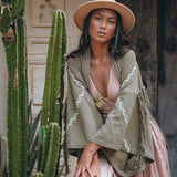 A woman in a chic ensemble leans against a tall cactus, donning a wide-brimmed hat and the Sage Colour Organic Cotton Poncho Cape by AYA Sacred Wear—a green jacket with fringes and shamanic embroidery over a pink dress. In the backdrop, an ornate wooden door with decorative patterns completes the scene.