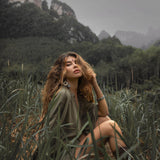 A woman with wavy hair in a sage green, hand-knotted romper sits in tall grass with a mountainous landscape in the background.