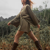 A person walks through a lush, mountainous landscape wearing a sage green hand-knotted jumpsuit from AYA Sacred Wear and brown boots. The sky is overcast, and the background features dense greenery and tall, rocky mountains. The individual's hair flows freely as they move in a comfortable summer style.