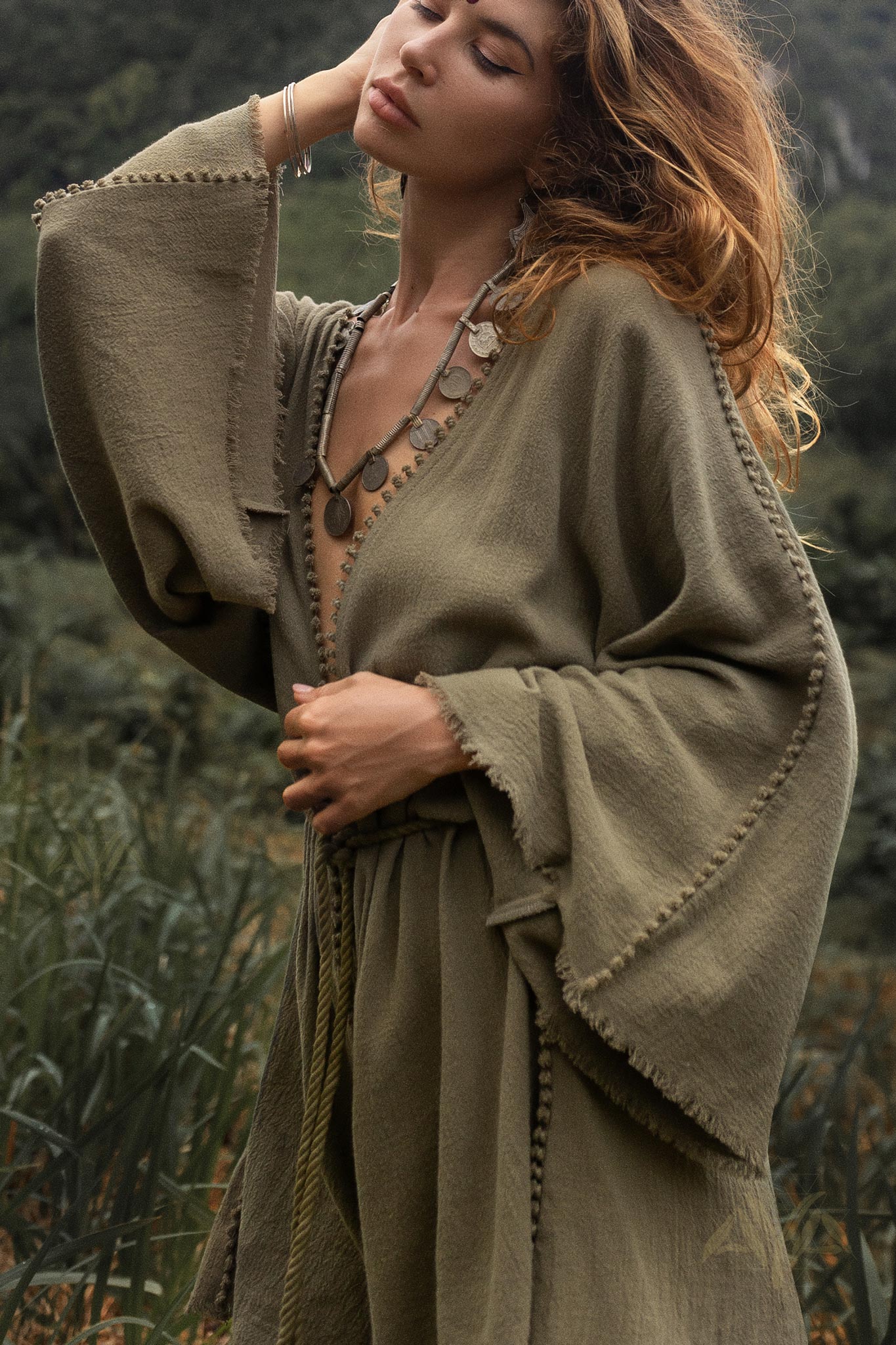 A woman with tousled hair poses in a sage green hand-knotted jumpsuit, featuring bell sleeves and intricate detailing. She holds one hand to her forehead and the other gently touches her waist. The background consists of lush, green vegetation, creating a serene, natural atmosphere.