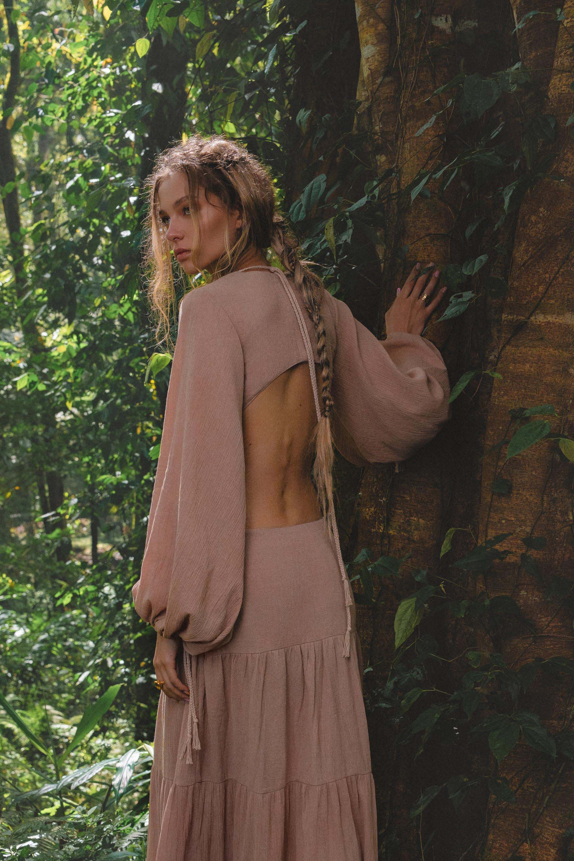 A woman with long, wavy, braided hair stands in a lush forest wearing the AYA Sacred Wear Samudra Dress in Blush Rose—a backless boho maxi. She glances over her shoulder, hand on a vine-draped tree trunk, brass spiral bracelet on her wrist.