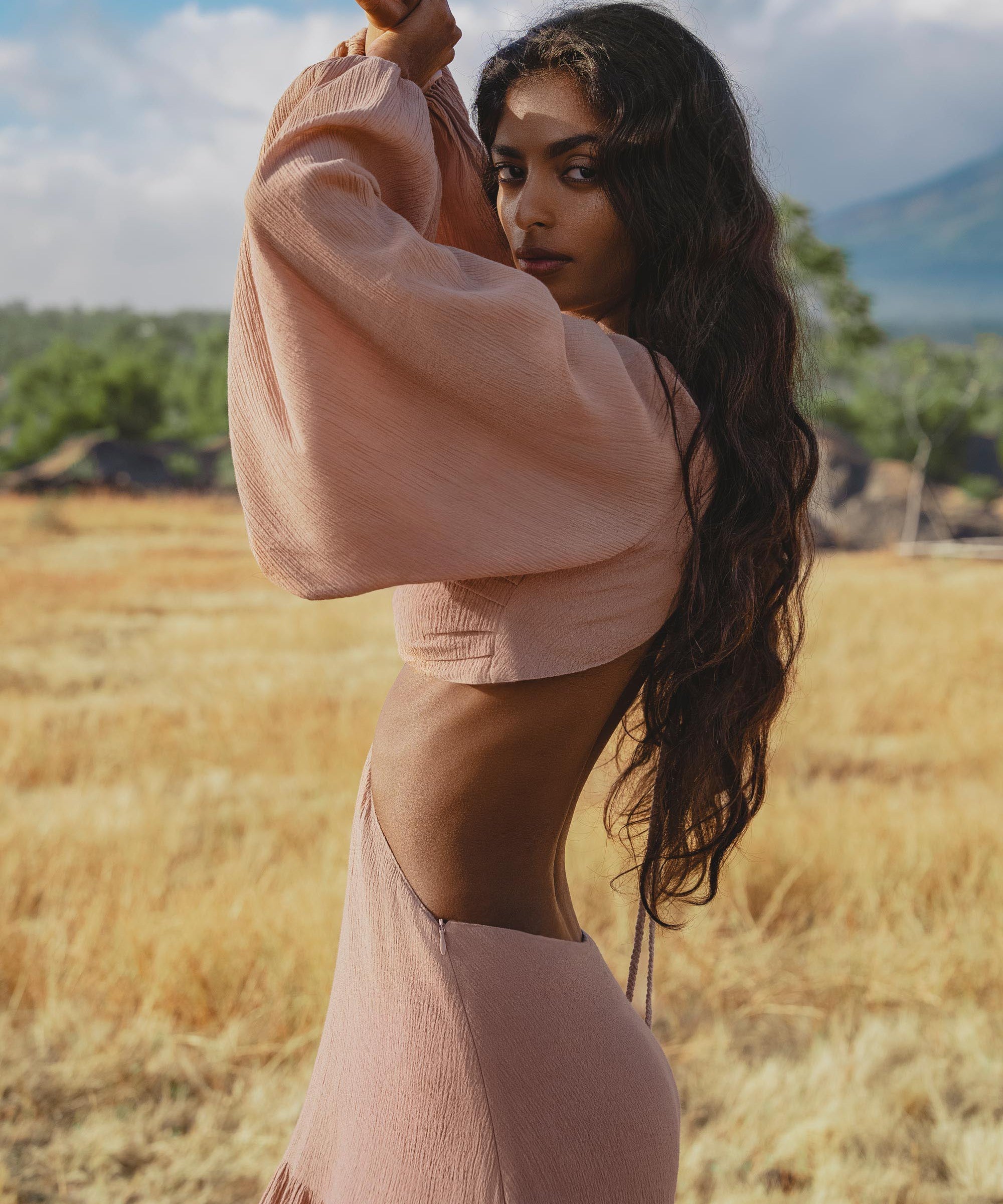 A woman with long wavy hair poses in a dry, grassy field wearing the AYA Sacred Wear Samudra Dress in Blush Rose—a backless boho maxi dress—paired with a handmade brass spiral. Green trees and distant mountains frame the scene.