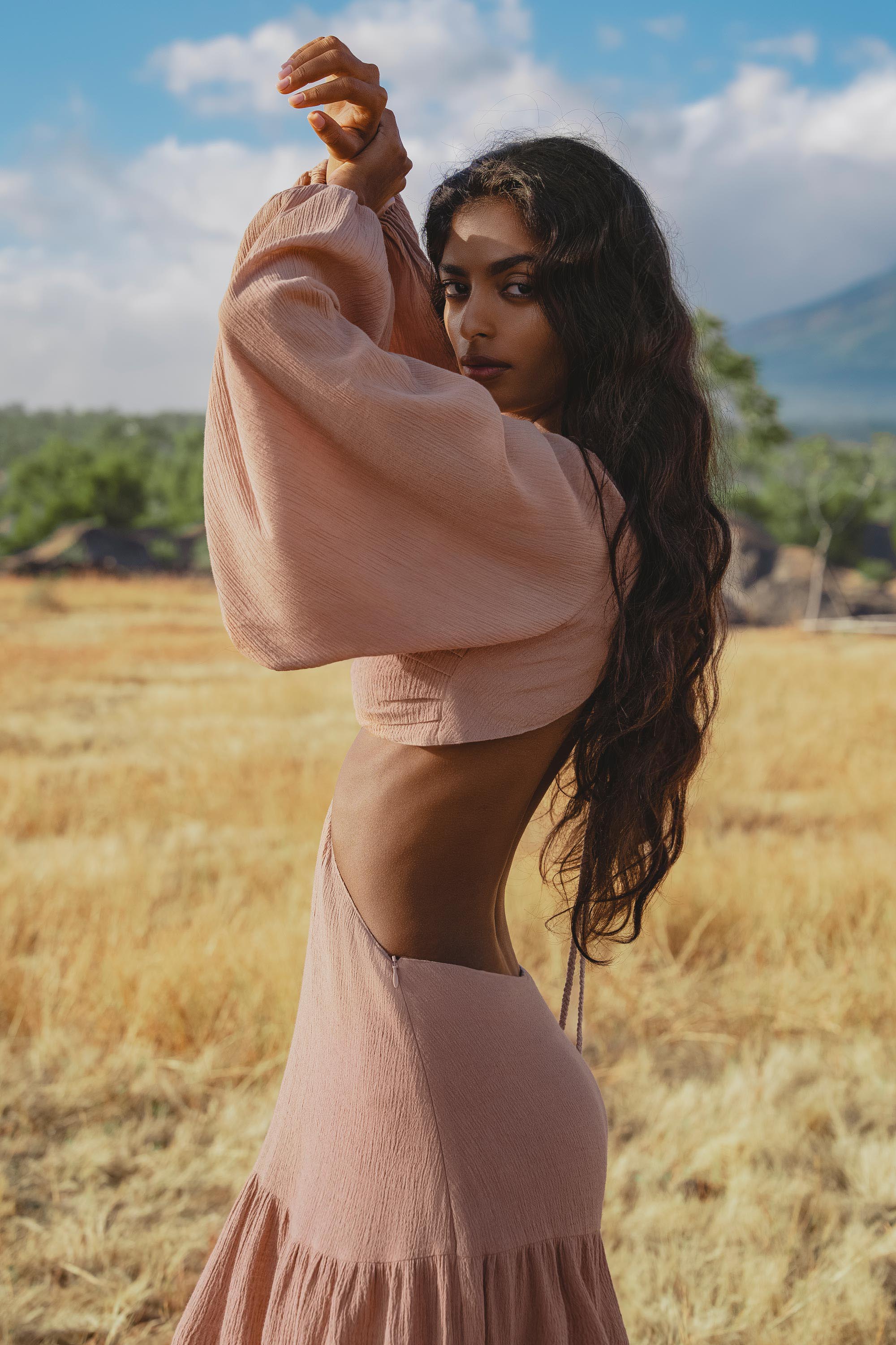 A woman with long wavy hair poses in a dry, grassy field wearing the AYA Sacred Wear Samudra Dress in Blush Rose—a backless boho maxi dress—paired with a handmade brass spiral. Green trees and distant mountains frame the scene.