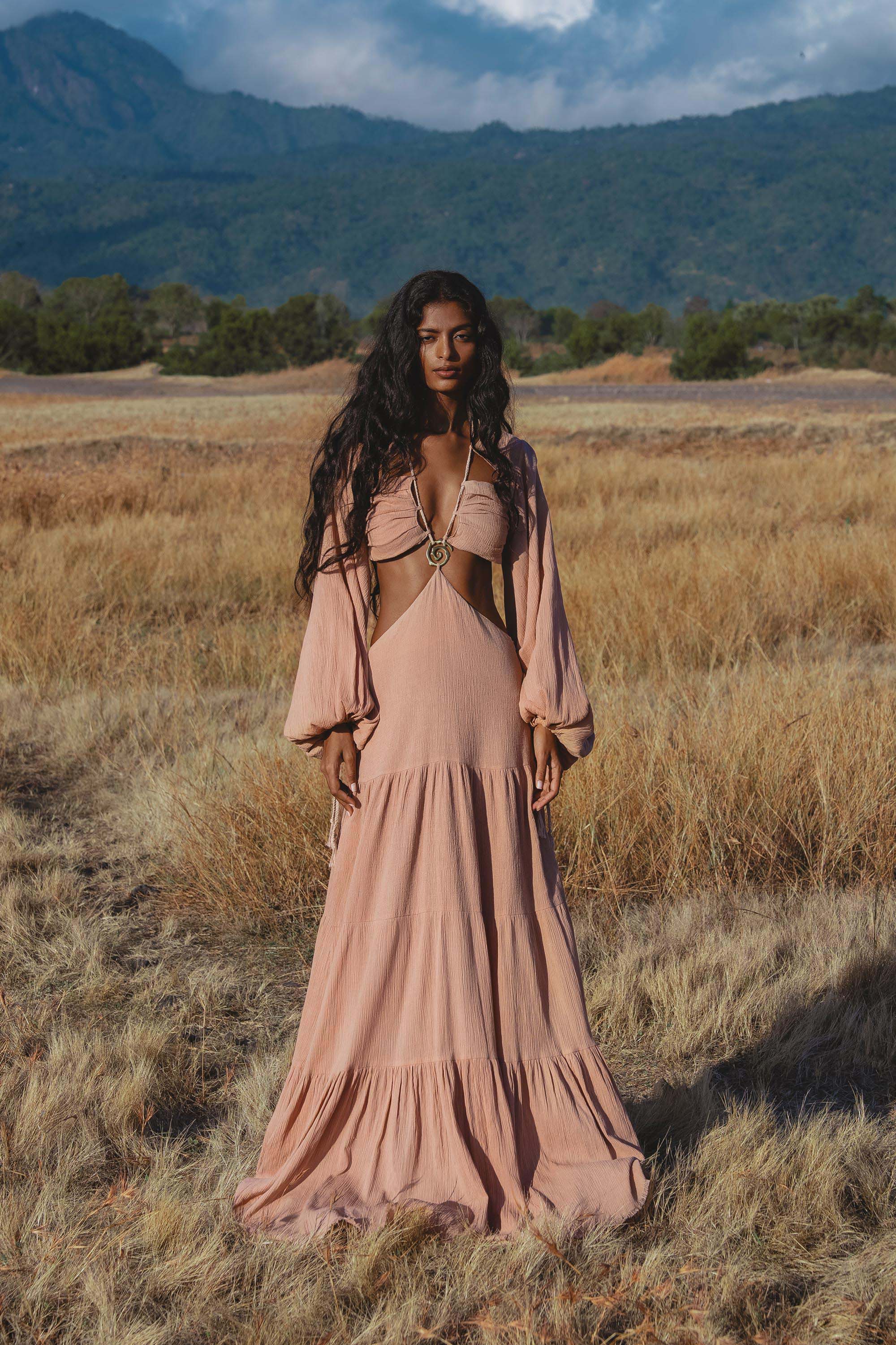 A woman with long, wavy hair stands in a dry grassy field wearing the AYA Sacred Wear Samudra Dress in Blush Rose—a backless boho maxi dress. Mountains and trees rise in the background beneath a partly cloudy sky.