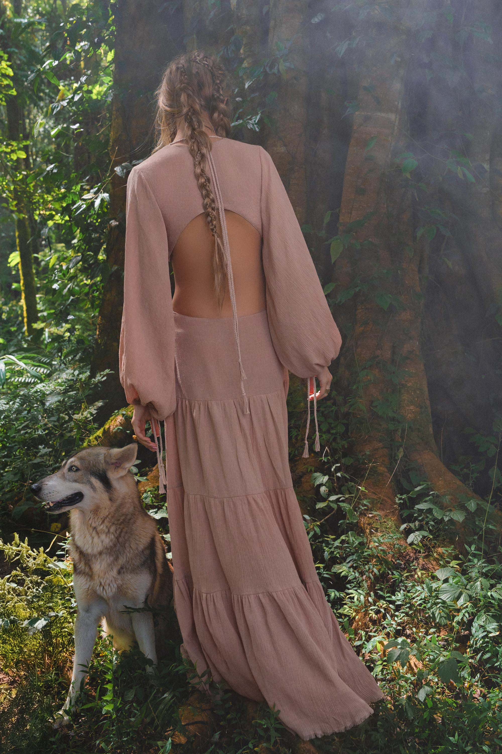 A woman with long braided hair stands in a lush forest, wearing the flowing Samudra Dress in Blush Rose by AYA Sacred Wear. Beside her, a wolf-like dog faces away, both framed by greenery and a large tree trunk.