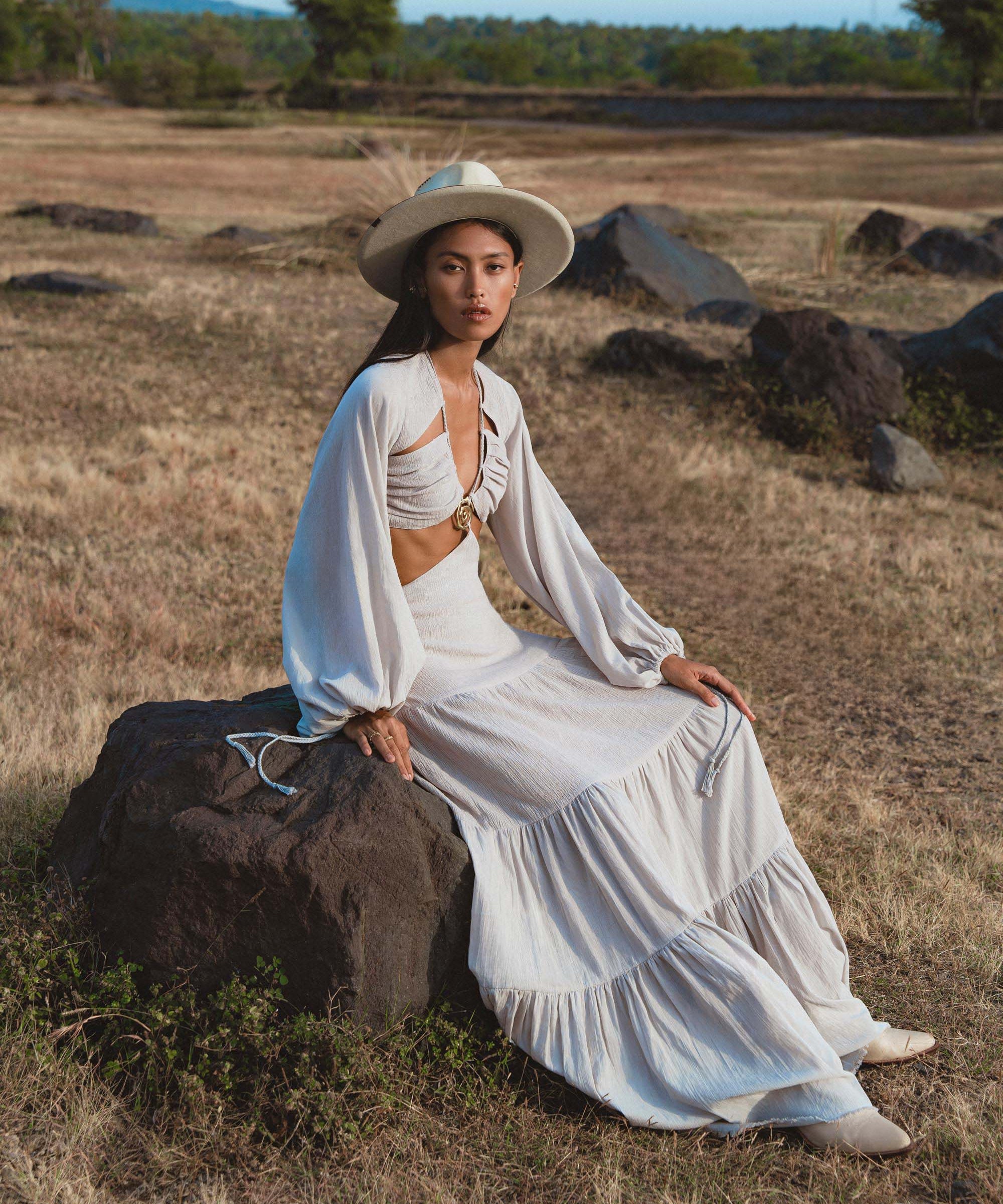 A woman in a wide-brimmed hat and the AYA Sacred Wear Samudra Dress in Celestial Mist Blue sits on a rock among dry grass, her organic bohemian garment's billowy sleeves catching the light as she gazes at the camera.