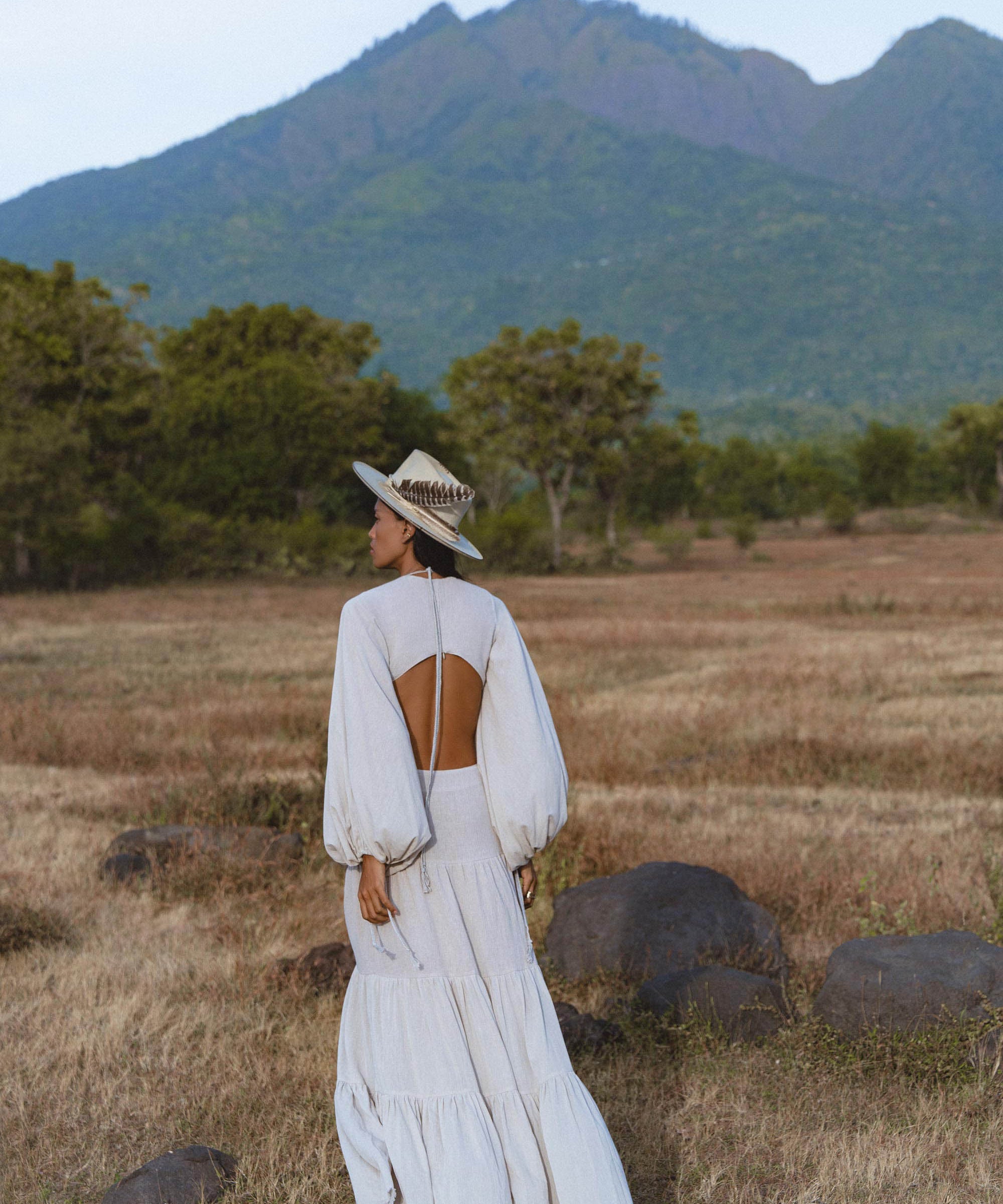 Wearing the AYA Sacred Wear Samudra Dress in Celestial Mist Blue—an organic, bohemian linen-cotton dress with an open back—a person stands in a dry grassy field, surrounded by green trees, rocks, and distant mountains beneath a cloudy sky.