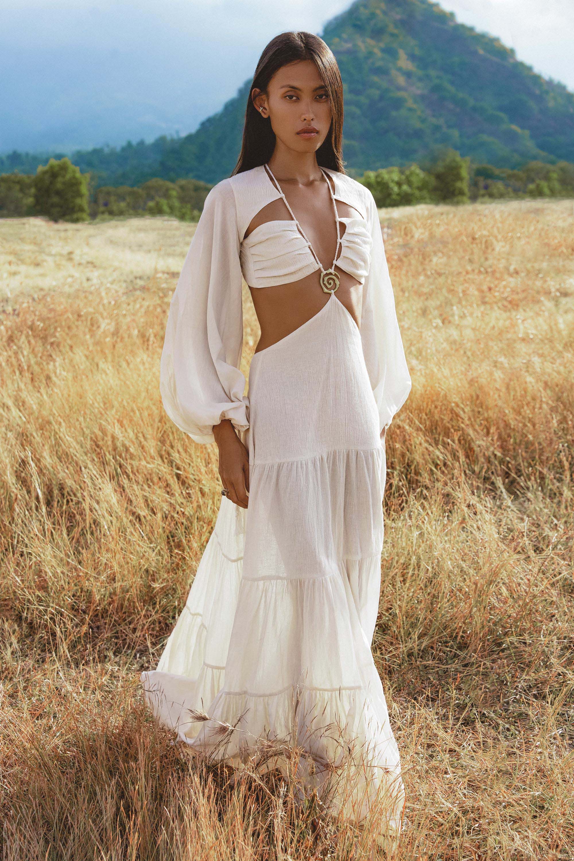 A woman with straight dark hair, calm expression, stands in a dry grass field wearing the Samudra Dress in Ivory Colour by AYA Sacred Wear—an organic bohemian linen-cotton dress with long sleeves, side cut-outs, and brass spiral chest detail. Green hills and mountains behind.