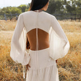 A woman stands in a sunny field, wearing the Samudra Dress in Ivory by AYA Sacred Wear—an organic bohemian dress with sheer sleeves, an open back tied with thin strings, and elegant linen cotton fabric. Her dark hair flows as the background blurs softly.