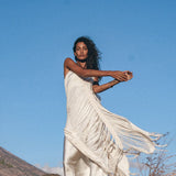 A woman poses against a clear blue sky, wearing the See Through Dress • Long Maxi Dress • Tribal Raw Cotton Cover-up by AYA Sacred Wear. She moves gracefully, with one arm raised and the other extended, amid dry grass and rocks.