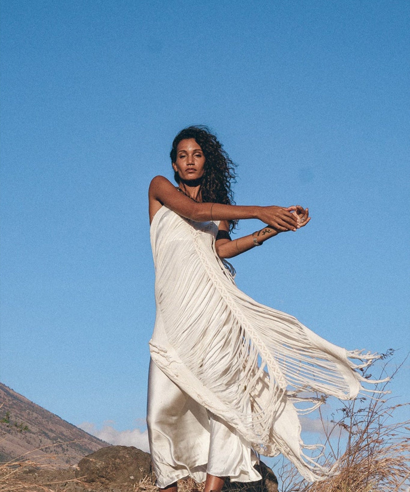 A woman poses against a clear blue sky, wearing the See Through Dress • Long Maxi Dress • Tribal Raw Cotton Cover-up by AYA Sacred Wear. She moves gracefully, with one arm raised and the other extended, amid dry grass and rocks.