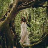 A woman in an AYA Sacred Wear See Through Dress, a long maxi dress made of tribal raw cotton, dances gracefully in a lush green forest, surrounded by twisting tree roots and foliage. Her arms are raised, and the scene is serene and mystical.