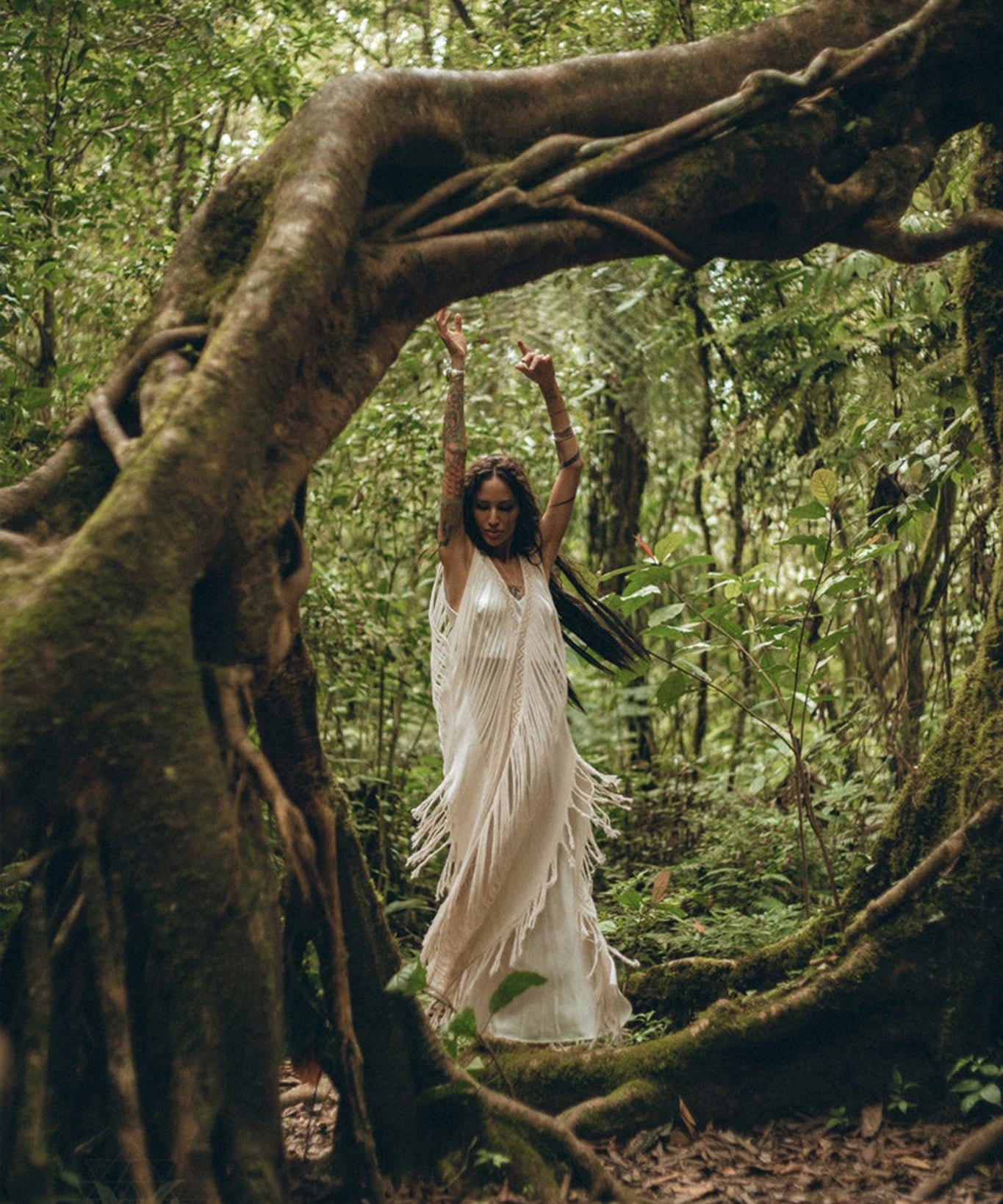 A woman in an AYA Sacred Wear See Through Dress, a long maxi dress made of tribal raw cotton, dances gracefully in a lush green forest, surrounded by twisting tree roots and foliage. Her arms are raised, and the scene is serene and mystical.