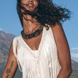 A woman with curly hair poses outdoors in a See Through Dress by AYA Sacred Wear, a long maxi dress designed as a tribal raw cotton cover-up. The mountain and blue sky form the backdrop as her tattooed arm, adorned with a bangle and a statement necklace, completes the look.