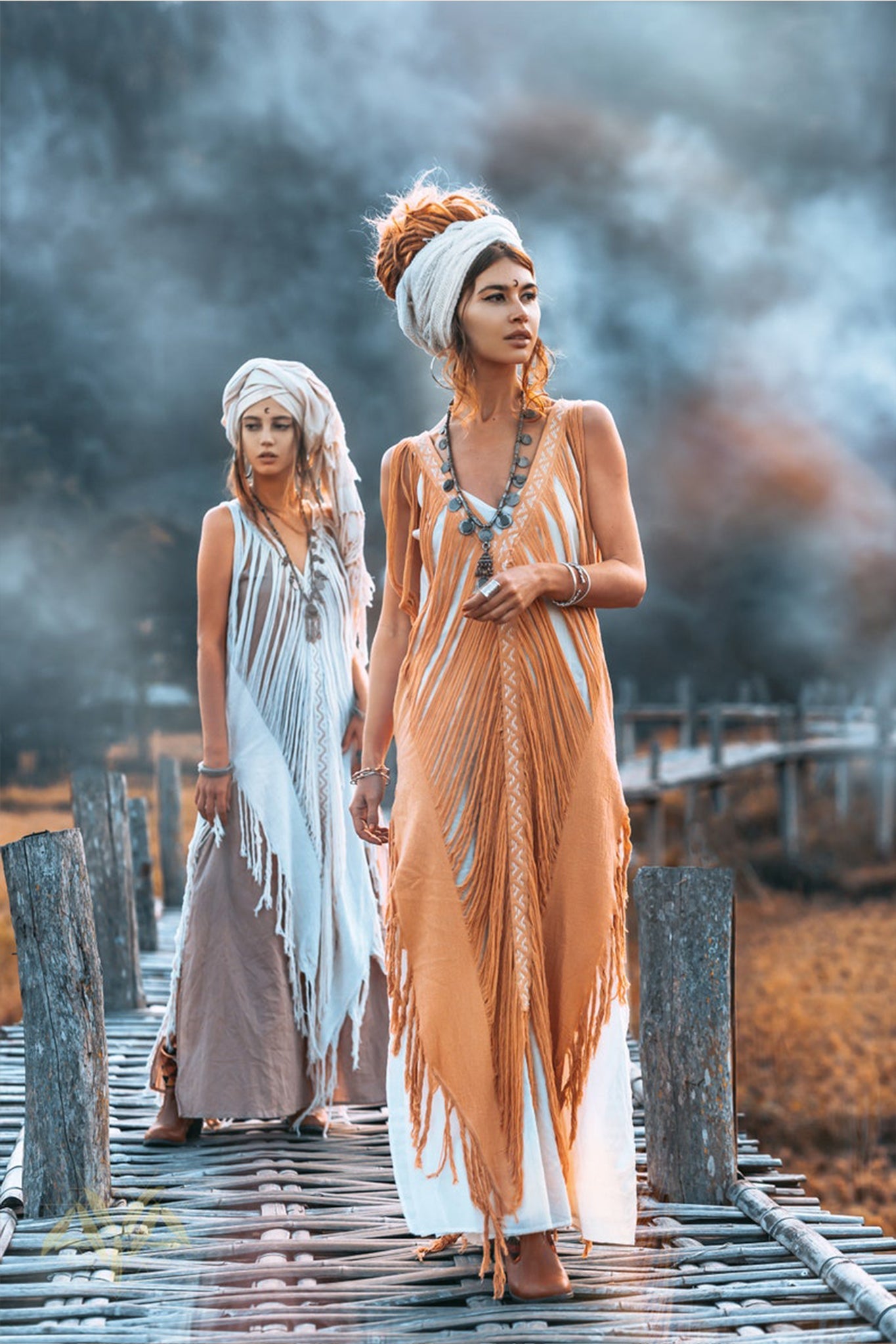 Two women with styled hair, dressed in AYA Sacred Wear's See Through Dress – a long maxi tribal raw cotton cover-up adorned with handmade embroidery and jewelry, stand on a rustic wooden walkway in a foggy, natural setting. One is in the foreground, the other in the background, both showcasing a bohemian style.