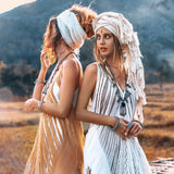 Two women wearing AYA Sacred Wear's Tribal Raw Cotton Cover-ups stand back to back in a field at sunset. One faces left, the other right, with majestic mountains as their backdrop. Their bohemian style is accentuated by layered necklaces and bracelets, complemented by the handmade embroidery of their See Through Long Maxi Dresses and head wraps.