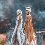 Two women pose on a wooden path, adorned in fringed, earth-toned See Through Dresses from AYA Sacred Wear's Long Maxi Dress collection. Each dress features exquisite handmade embroidery paired with elegant headwraps. They glance back at the camera amidst an atmospheric, misty backdrop that enhances the mystical ambiance.