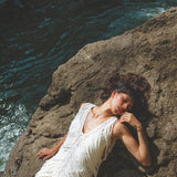 Dressed in AYA Sacred Wear's See Through Dress • Long Maxi Dress • Tribal Raw Cotton Cover-up, a woman reclines on rocks by the river. Sunlight dances across the textured fabric, while the nearby flowing water shimmers. Her dark hair cascades around her, and she exudes relaxation and serenity.