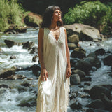 Standing atop a rock by a flowing river, an individual is elegantly dressed in the AYA Sacred Wear's See Through Dress – a Long Maxi Dress in Tribal Raw Cotton. The intricate handmade embroidery adds charm to the sleeveless white cover-up, set against a backdrop of lush greenery illuminated by natural daylight.