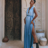 A person stands confidently in a Sky Blue Ombre Dyed Dress from AYA Sacred Wear, showcasing its boho elegance with a high slit, while posing against a textured wall with rustic wooden doors. They complement the look with black ankle boots and bracelets, as clay pots are artfully placed on the ground nearby.