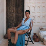 A person wearing a Sky Blue Ombre Dyed Dress from AYA Sacred Wear, complemented by black boots, sits on a wooden chair in front of an intricately carved wooden door. The scene is enhanced by pottery and textured beige walls with ornate architectural details.