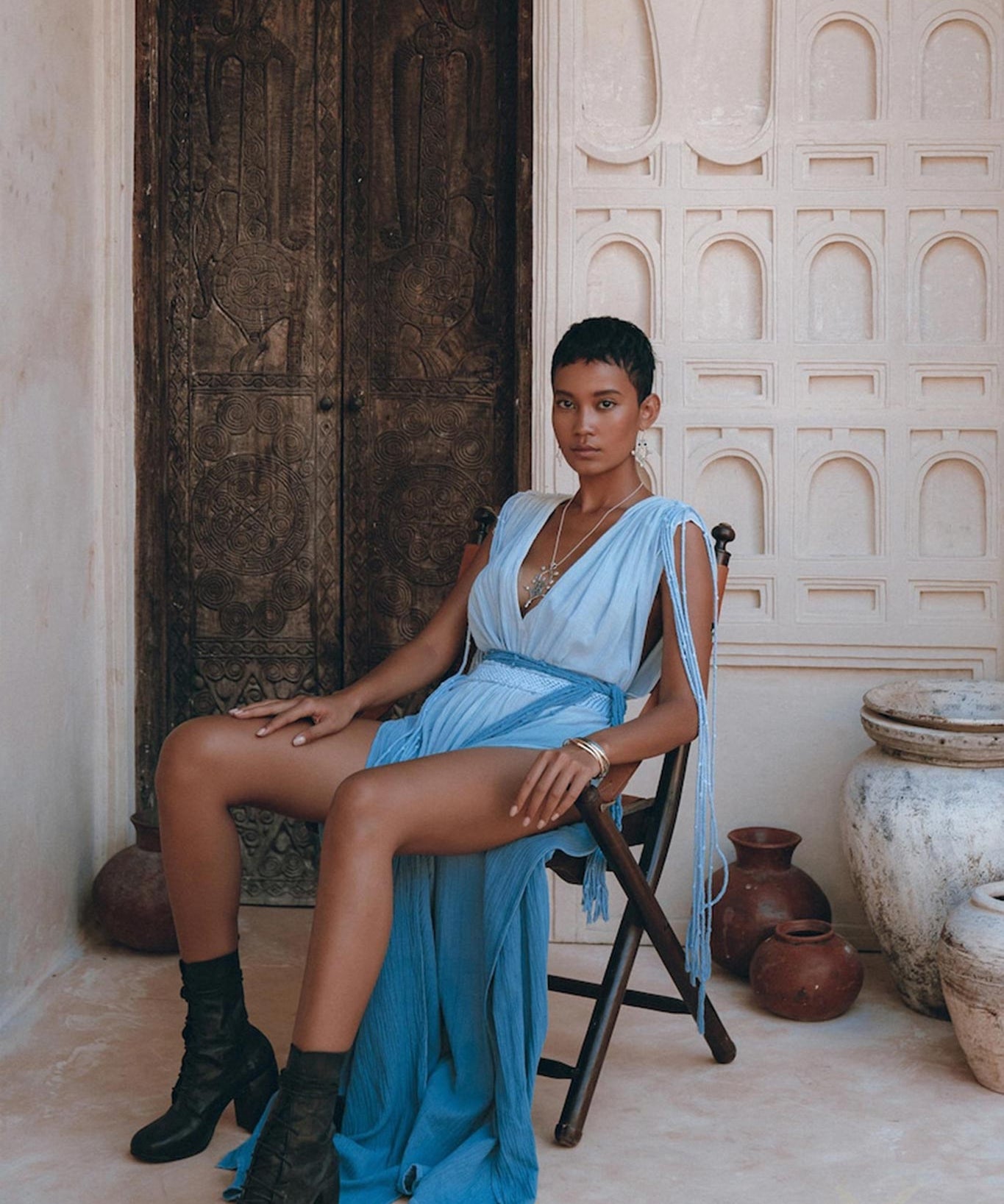 A person wearing a Sky Blue Ombre Dyed Dress from AYA Sacred Wear, complemented by black boots, sits on a wooden chair in front of an intricately carved wooden door. The scene is enhanced by pottery and textured beige walls with ornate architectural details.