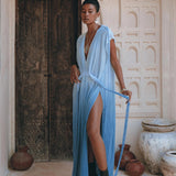 A person stands confidently in front of an ornate wooden door, wearing the AYA Sacred Wear Sky Blue Ombre Dyed Dress—a boho wedding dress with a flowing blue design and a thigh-high slit. They hold a rope-like belt and wear high-heeled boots. The background features rustic pots and light-colored walls with intricate designs.