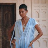 A person poses confidently in an AYA Sacred Wear creation, the Sky Blue Ombre Dyed Dress. This handcrafted boho wedding dress, complete with a deep V-neckline and high slits, elegantly boasts flowing fabric. They accessorize with a necklace and earrings, set against the backdrop of a decorative wall and an ornate wooden door.