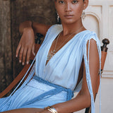 A person with short hair sits confidently in a wooden chair, adorned in an AYA Sacred Wear Sky Blue Ombre Dyed Dress, a flowing, sleeveless boho wedding dress complemented by silver jewelry. The handcrafted macrame design elegantly drapes across the ornate, textured background.