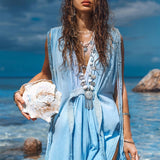 A woman stands on a beach holding a large seashell. Her long wet hair cascades over an AYA Sacred Wear Sky Blue Ombre Dyed Dress, a boho wedding dress adorned with handmade macrame details. The blue fabric of the dress mirrors the ocean's shades, its flowing design harmonizing beautifully with the serene backdrop of ocean and rocks under the cloudy sky.