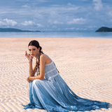 A woman sits on a sunlit beach in an AYA Sacred Wear Sky Blue Greek Goddess Dress, crafted from botanically dyed organic cotton. Her braided hair complements the partly cloudy sky and distant islands in the background as she gazes at the camera.