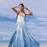 A woman stands barefoot on a sandy beach, wearing an AYA Sacred Wear Sky Blue Greek Goddess Dress botanically dyed in blue shades of organic cotton. Her braided hair and tribal bracelets sway in the gentle breeze, under a sky dotted with clouds as the ocean softly whispers nearby.